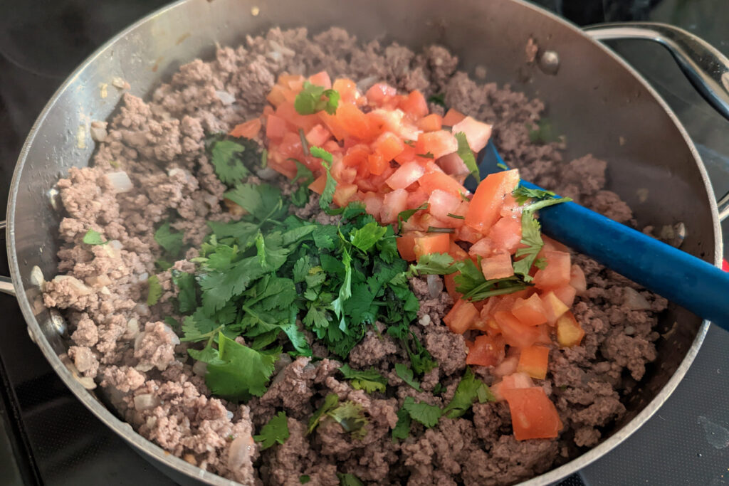 Cilantro and tomatoes cooking with ground beef in a pan.