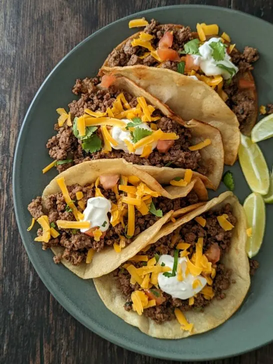 Four ground beef tacos topped with sour cream, cheddar cheese, and cilantro.