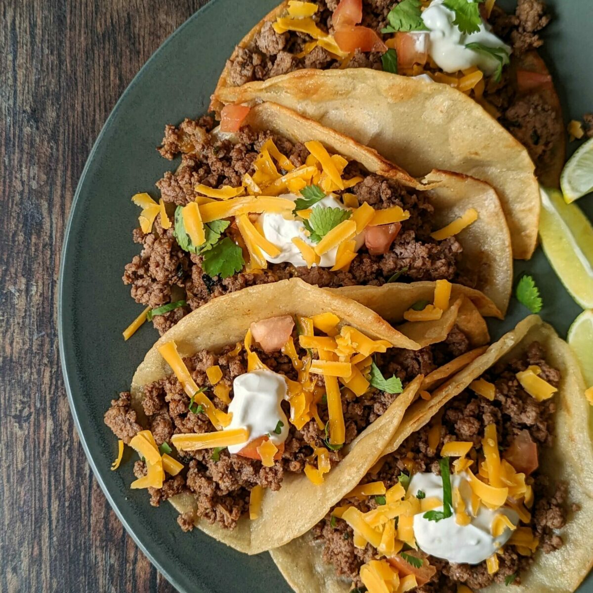 Four ground beef tacos topped with sour cream, cheddar cheese, and cilantro.