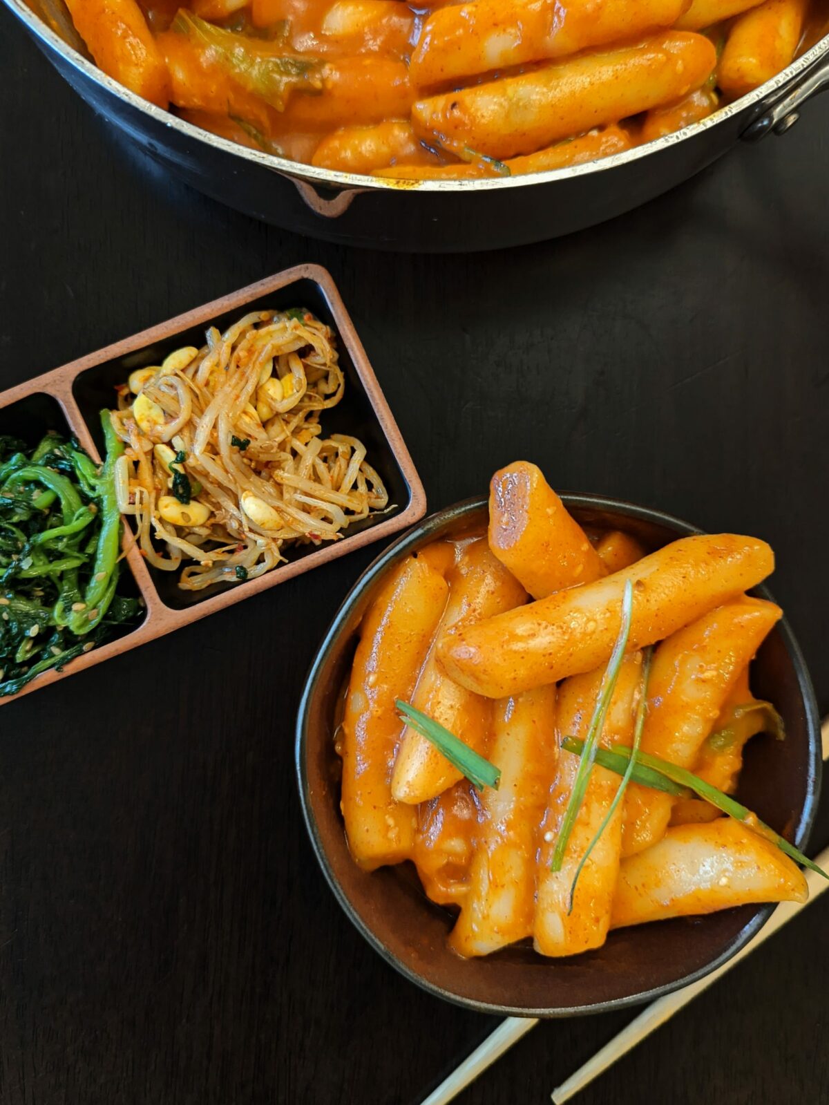 A small bowl of tteokbokki garnished with scallions and served with pickled sides.