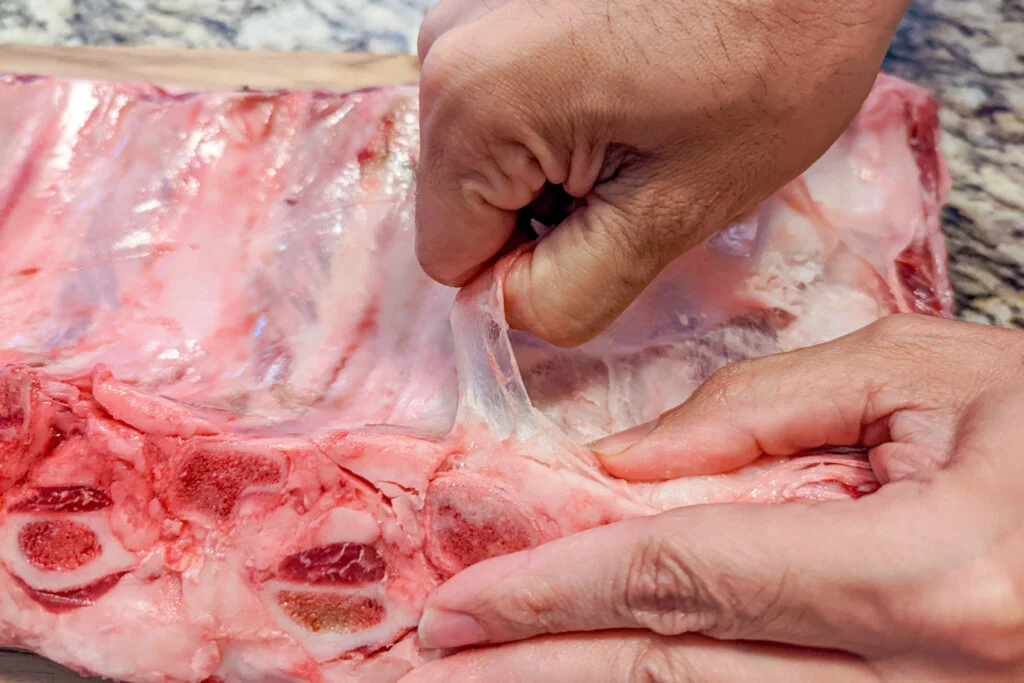 A hand peeling back the membrane from the beef ribs.