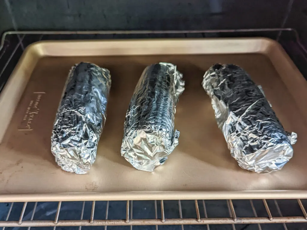 Corn on the cob wrapped in foil on a baking sheet.