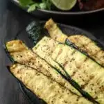 Grilled zucchini in a serving dish with grilled chicken in the background.