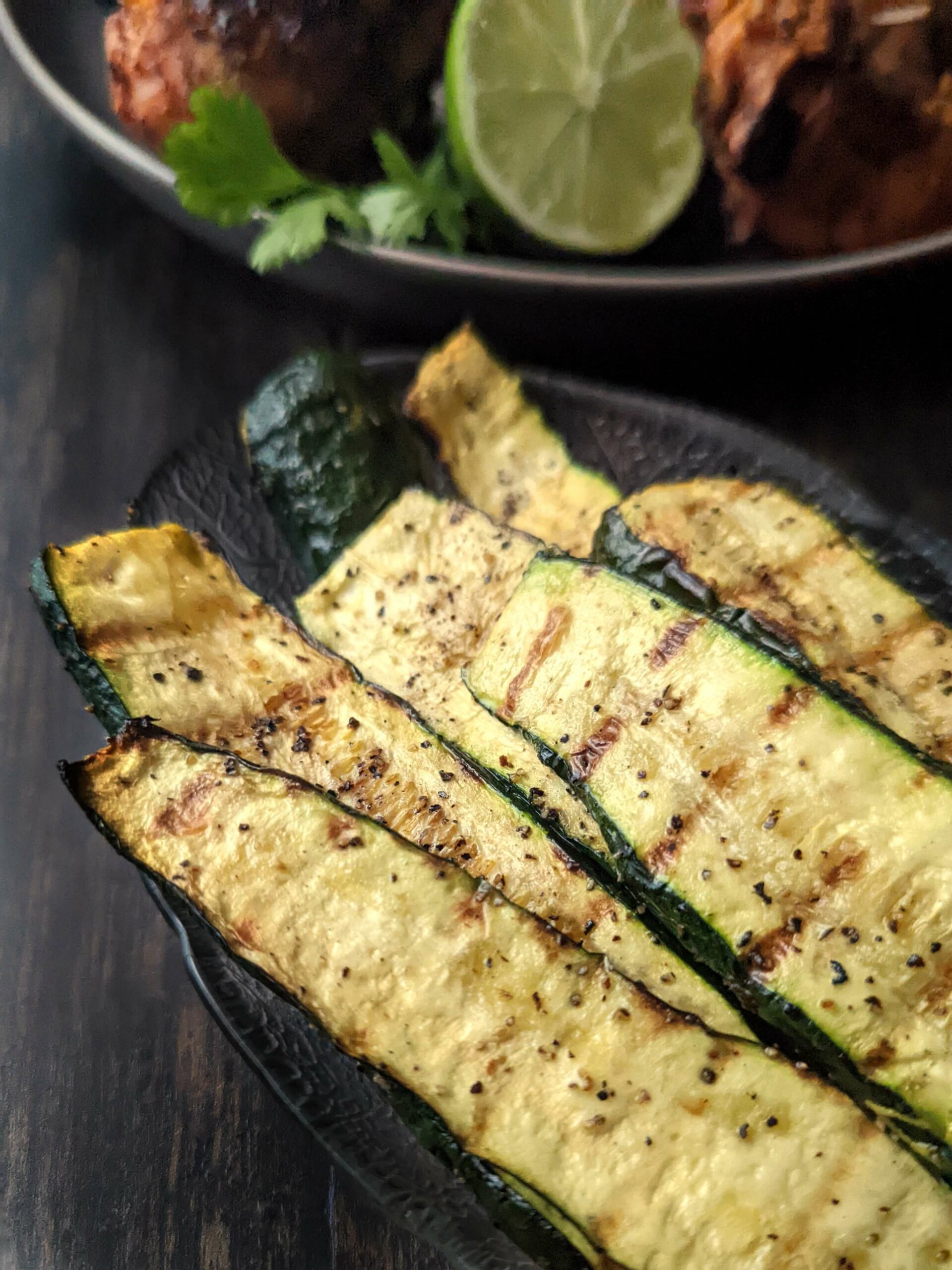 Grilled zucchini in a serving dish with grilled chicken in the background.