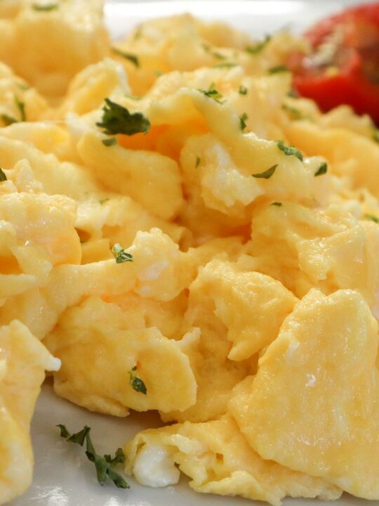 Scrambled eggs topped with chopped parsley with tomatoes in the background.