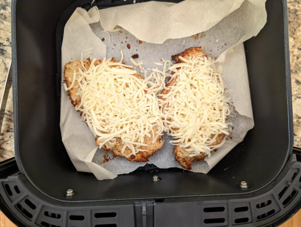 Two chicken breasts topped with cheese cooking in the air fryer.