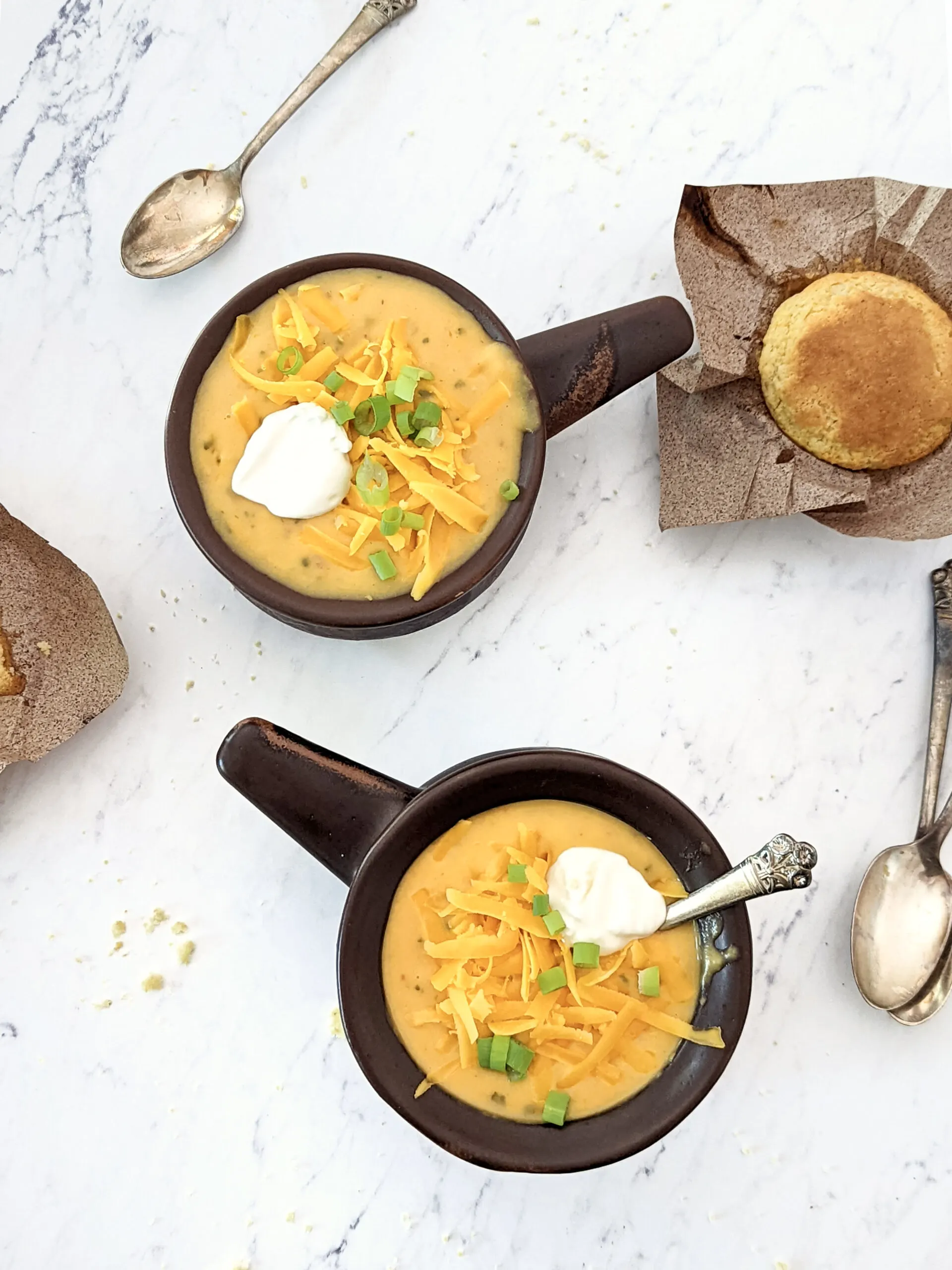 Two bowls of instant pot potato soup topped with green onion, cheddar cheese, and sour cream with corn muffins in the background.