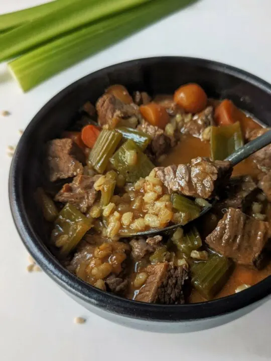 A bowl of beef and barley soup with celery and barley in the background.