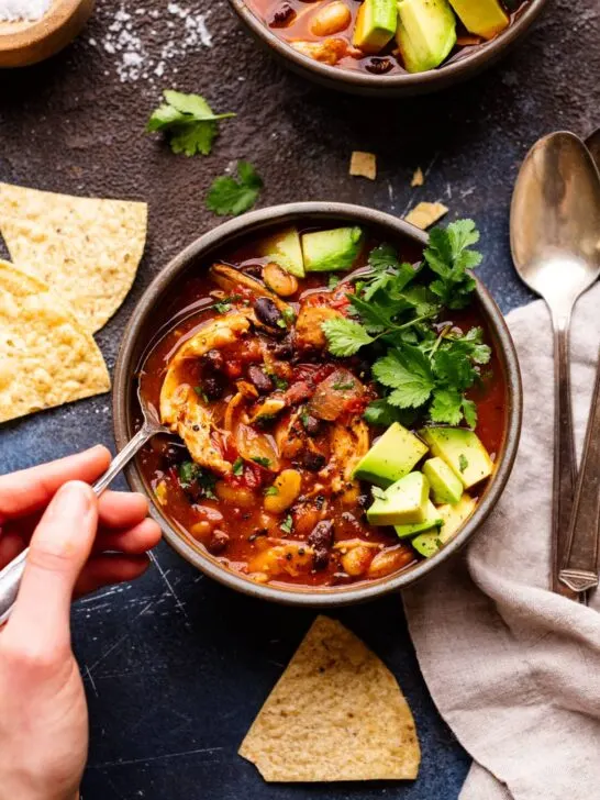 A hand holding a spoon and scooping out a spoonful of chicken chili topped with cilantro and avocado.