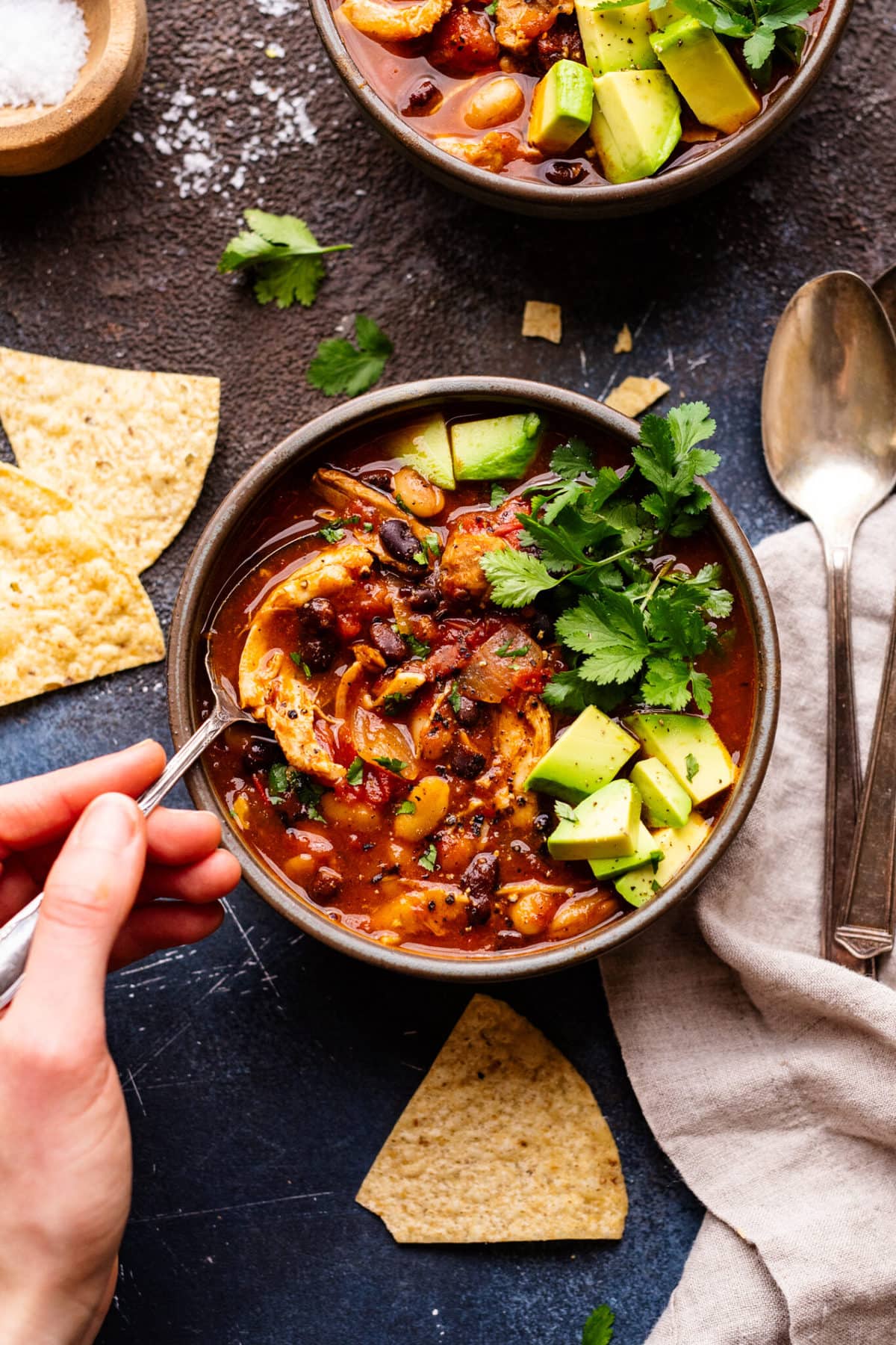 A hand holding a spoon and scooping out a spoonful of chicken chili topped with cilantro and avocado.