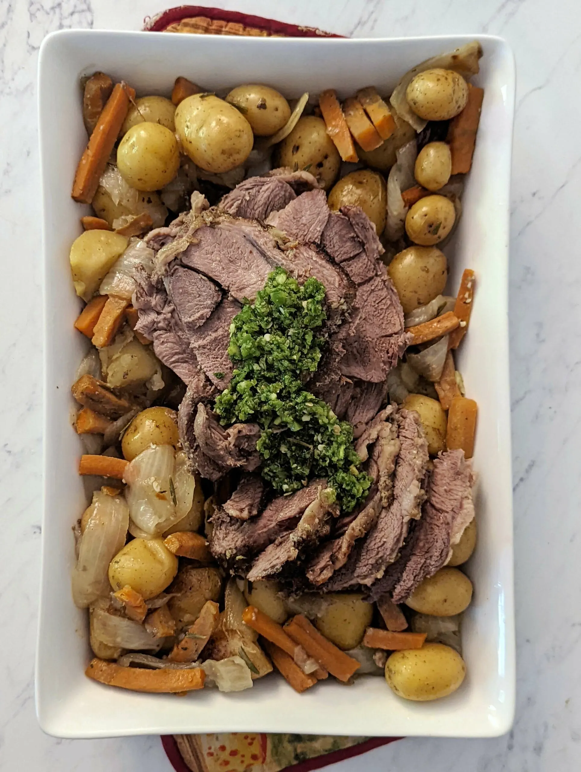 Sliced boneless leg of lamb roast in a serving dish with potatoes and carrots and garnished with skhug.
