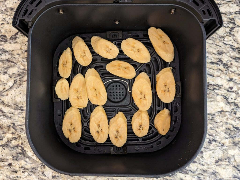 Uncooked plantain slices lined in the air fryer basket.