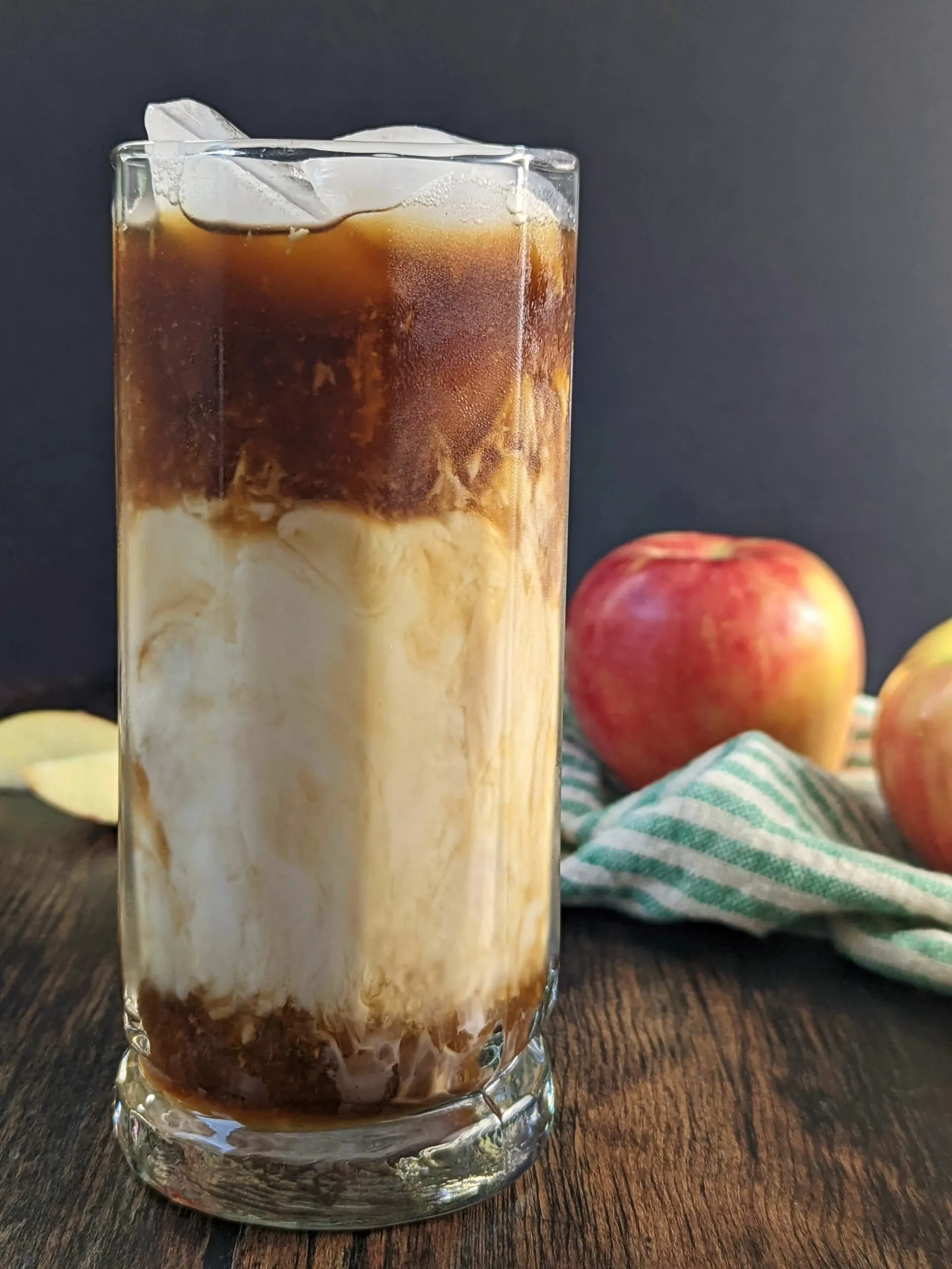 A glass of Iced Apple Crisp Macchiato Recipe with apples in the background.