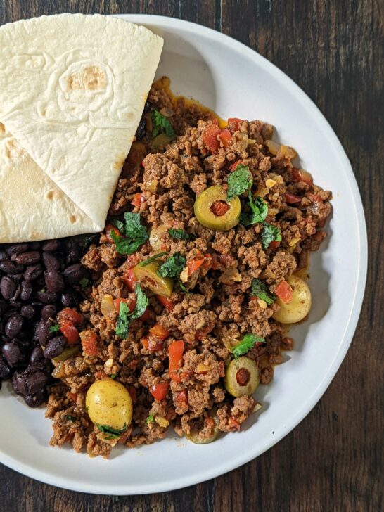 Beef picadillo on a plate black beans and a flour tortilla.