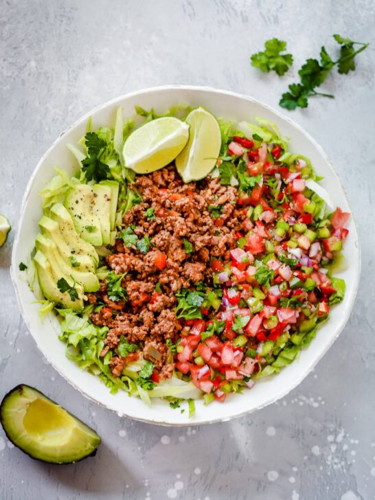 Ground beef taco salad in a bowl with avocado slices and lime wedges.