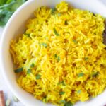 A bowl of turmeric yellow rice with cilantro in the background.
