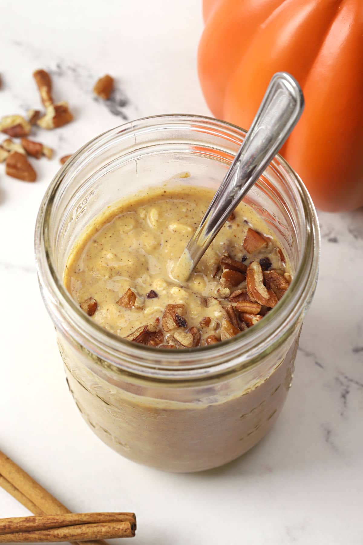 A jar of overnight oats topped with pecans and a pumpkin in the background.