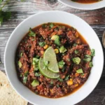 A bowl of chili topped with cilantro and lime wedges.