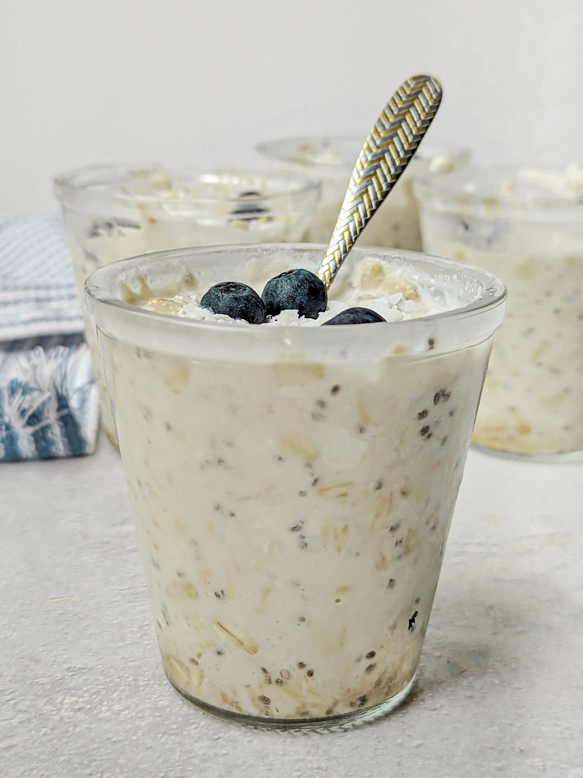 A close up of a container of overnight oats with coconut milk topped with blueberries and more containers in the background. 