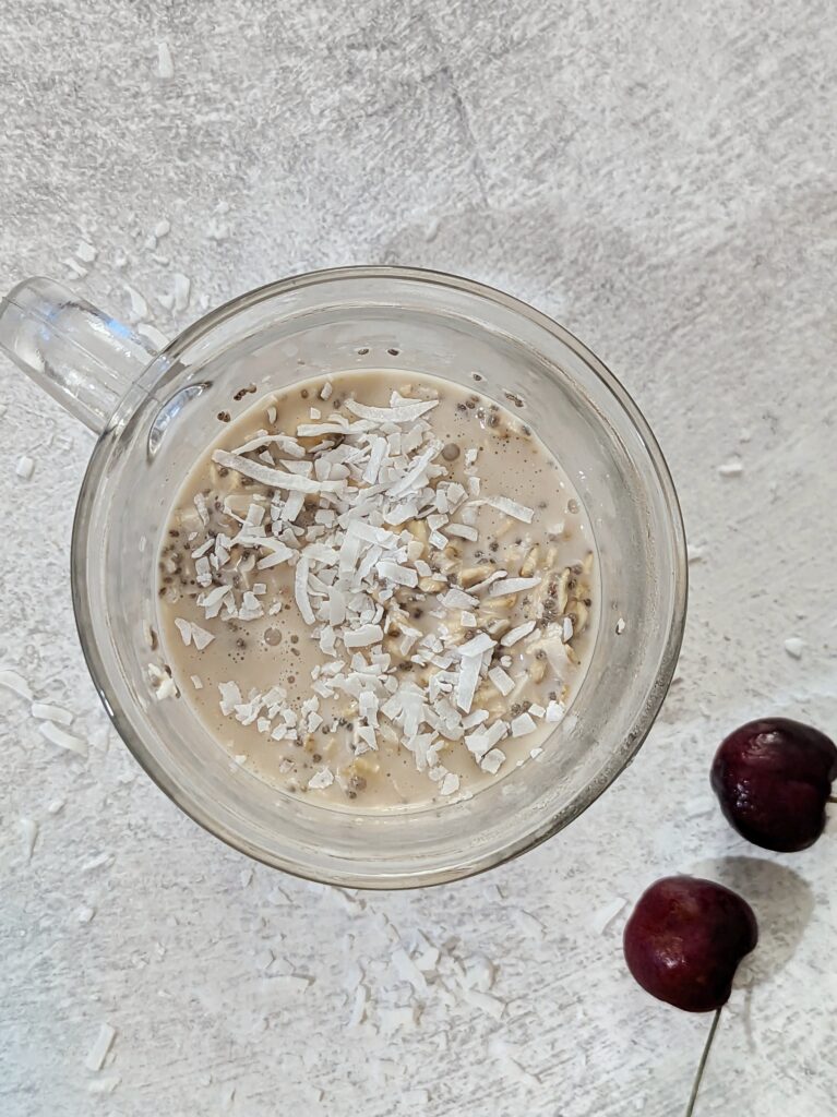 A cup over overnight oats topped with coconut flakes with cherries on the table.