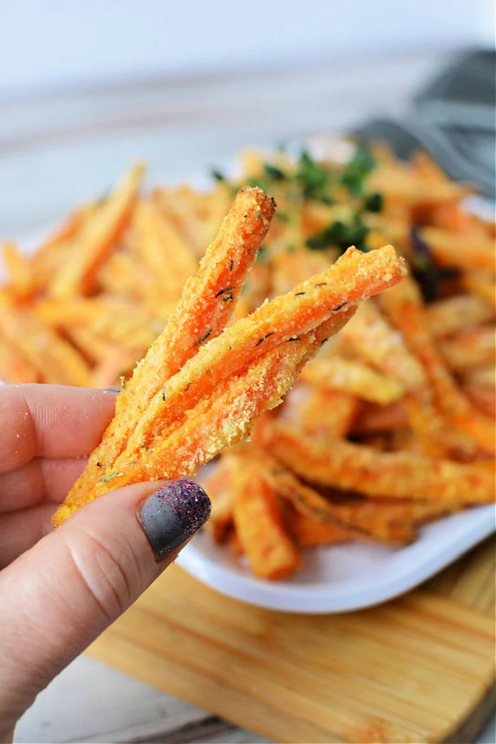A hand holding carrot fries over a plate of carrot fries.