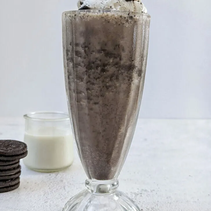 A close up of a milkshake without ice cream with whopped cream and an oreo on top with oreos in the background.