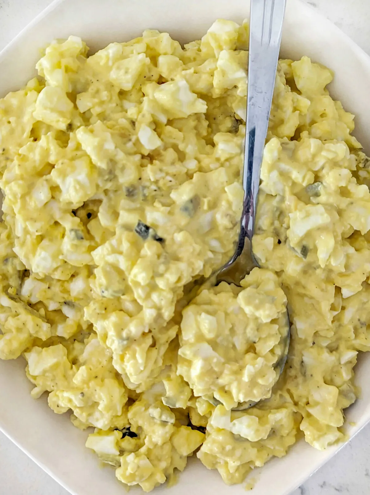 A close up of a bowl of egg salad with pickles with a spoon in it.