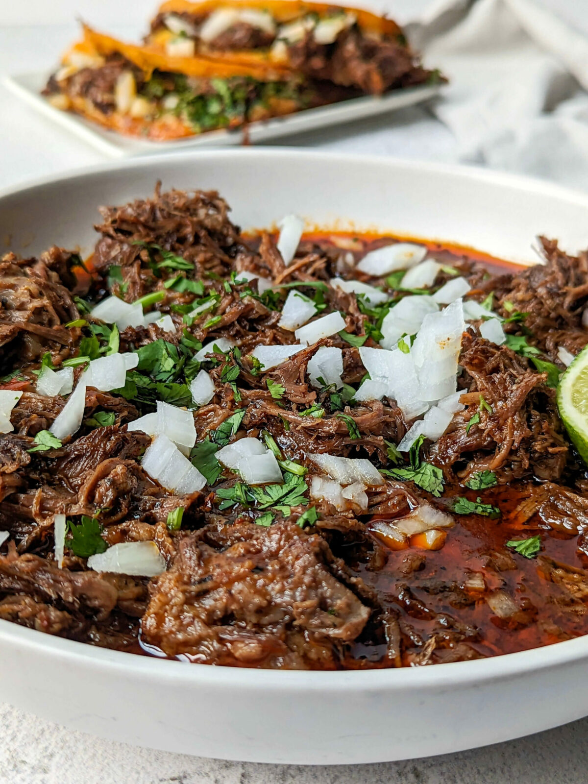 A bowl of birria con consome garnished with onion and cilantro with birria tacos in the background.