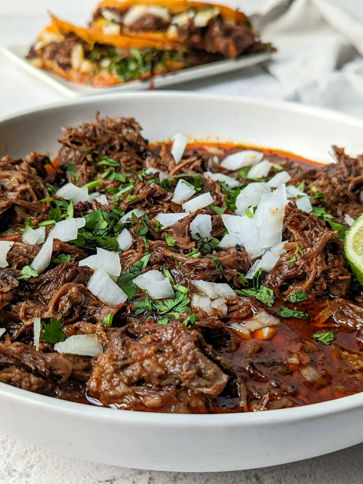 A bowl of birria con consome garnished with onion and cilantro with birria tacos in the background.