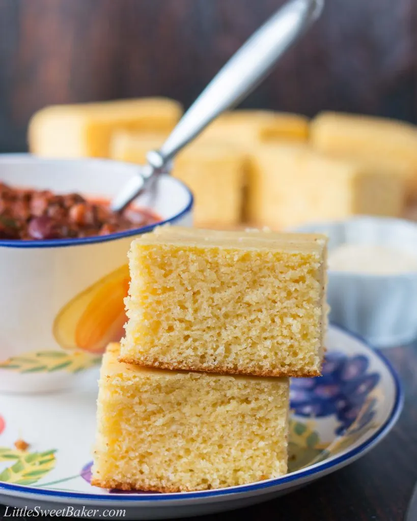 Two slices of cornbread stacked on a plate with a bowl of chili and more cornbread in the background.