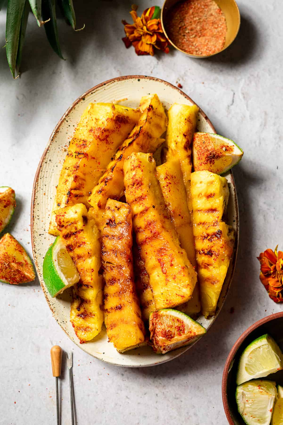 Grilled pineapple skewers are serving on a plate with lime wedges with chilies in the background.