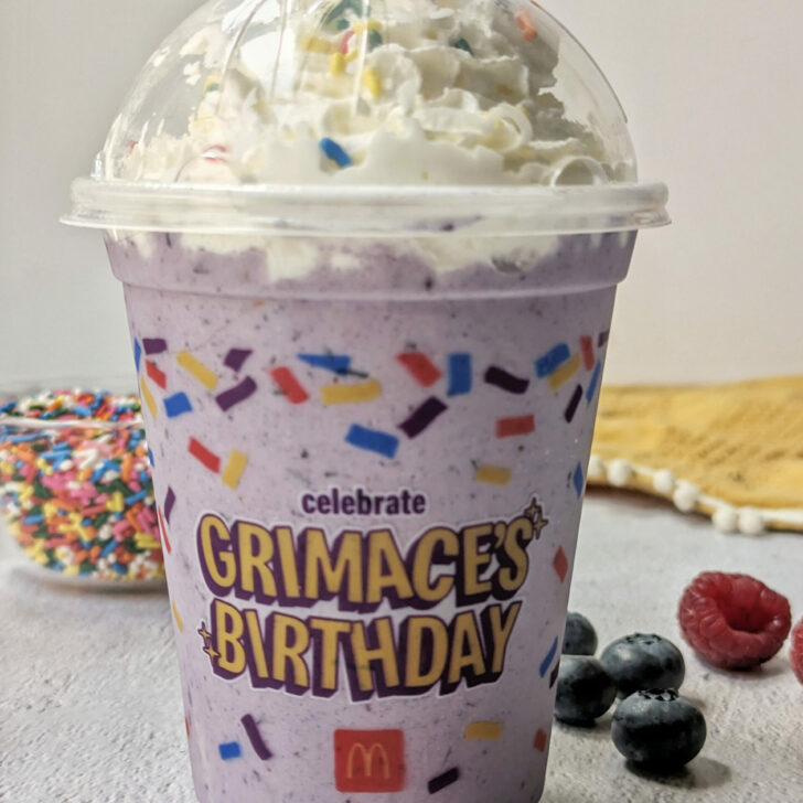 Grimace McDonalds Milkshake Recipe in a cup and topped with whipped cream and sprinkles, and surrounded by berries.