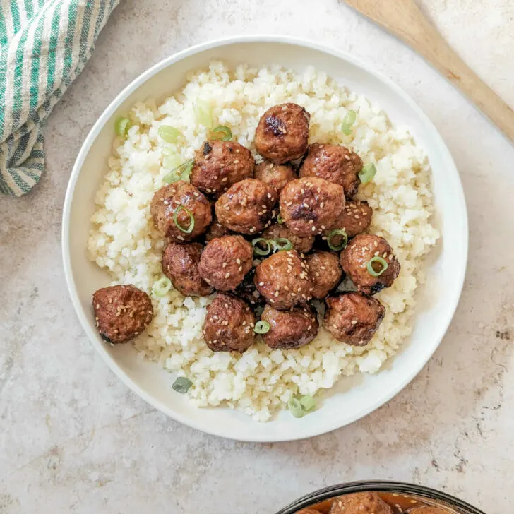 Instant Pot Frozen Meatballs served over cauliflower rice topped with scallions and a bowl of meatballs in the background.