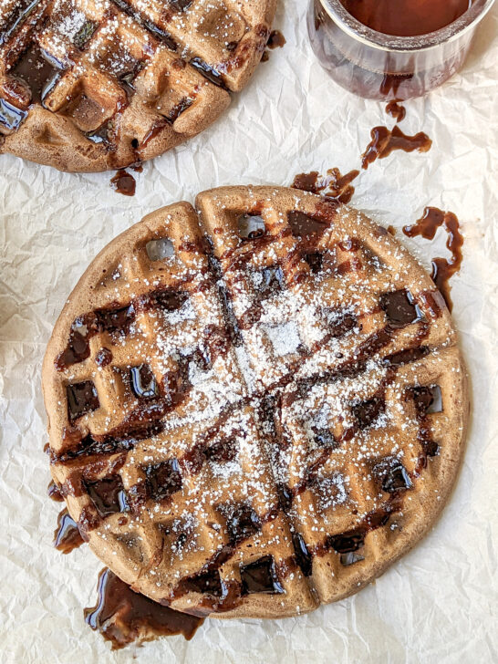 A nutella waffle topped with powdered sugar and chocolate sauce with another waffle in the background.