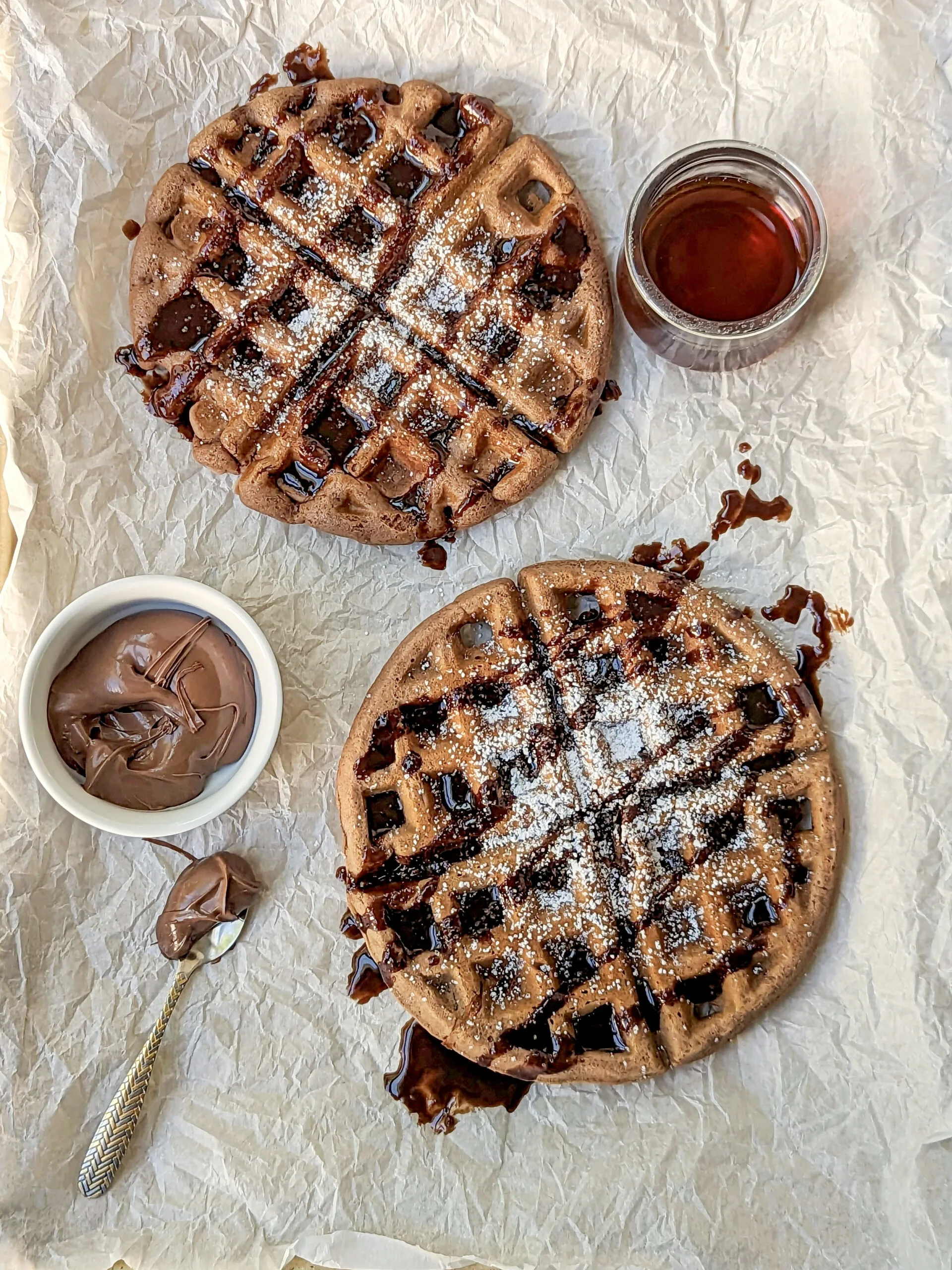 Two nutella waffles topped with powdered sugar and chocolate sauce with a small bowl of nutella and a small bowl of maple syrup.