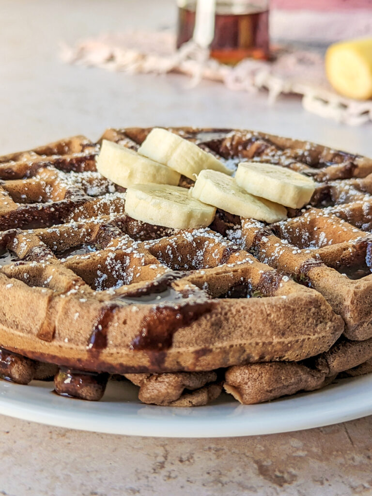 A nutella waffles topped with powdered sugar, chocolate sauce, and sliced bananas.