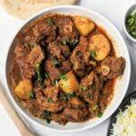Aloo gosht on a plate topped with chopped cilantro and rice and naan in the background.