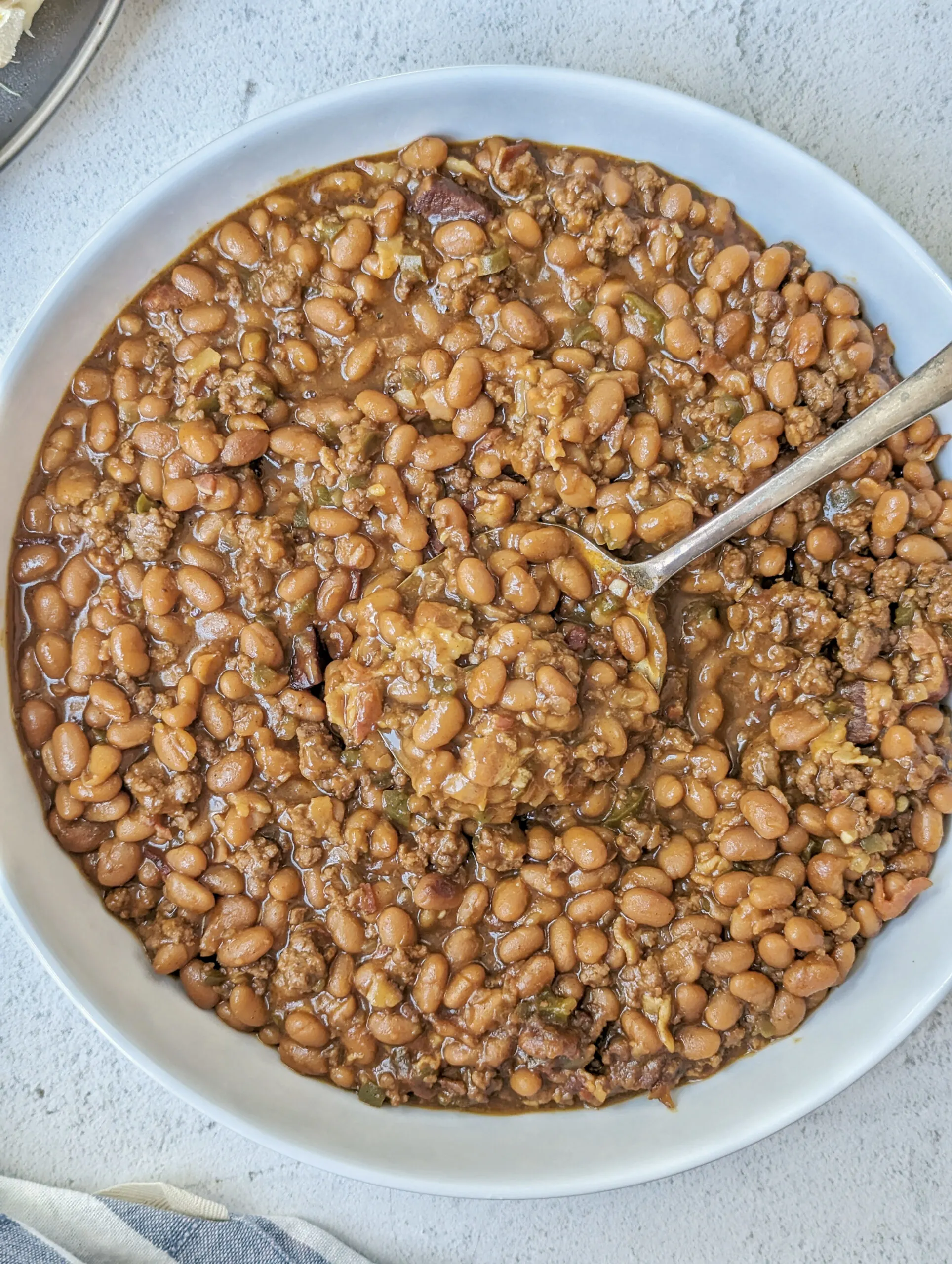 A serving bowl of baked beans with corn on the cob in the background.