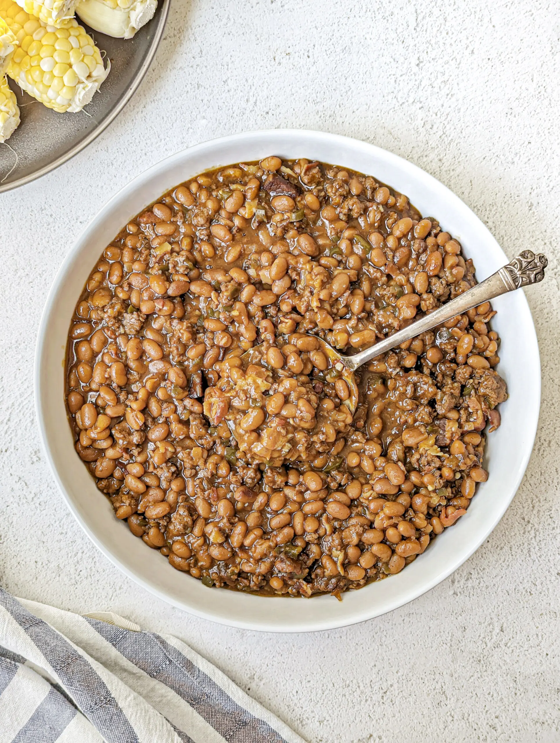 A serving bowl of baked beans with corn on the cob in the background.