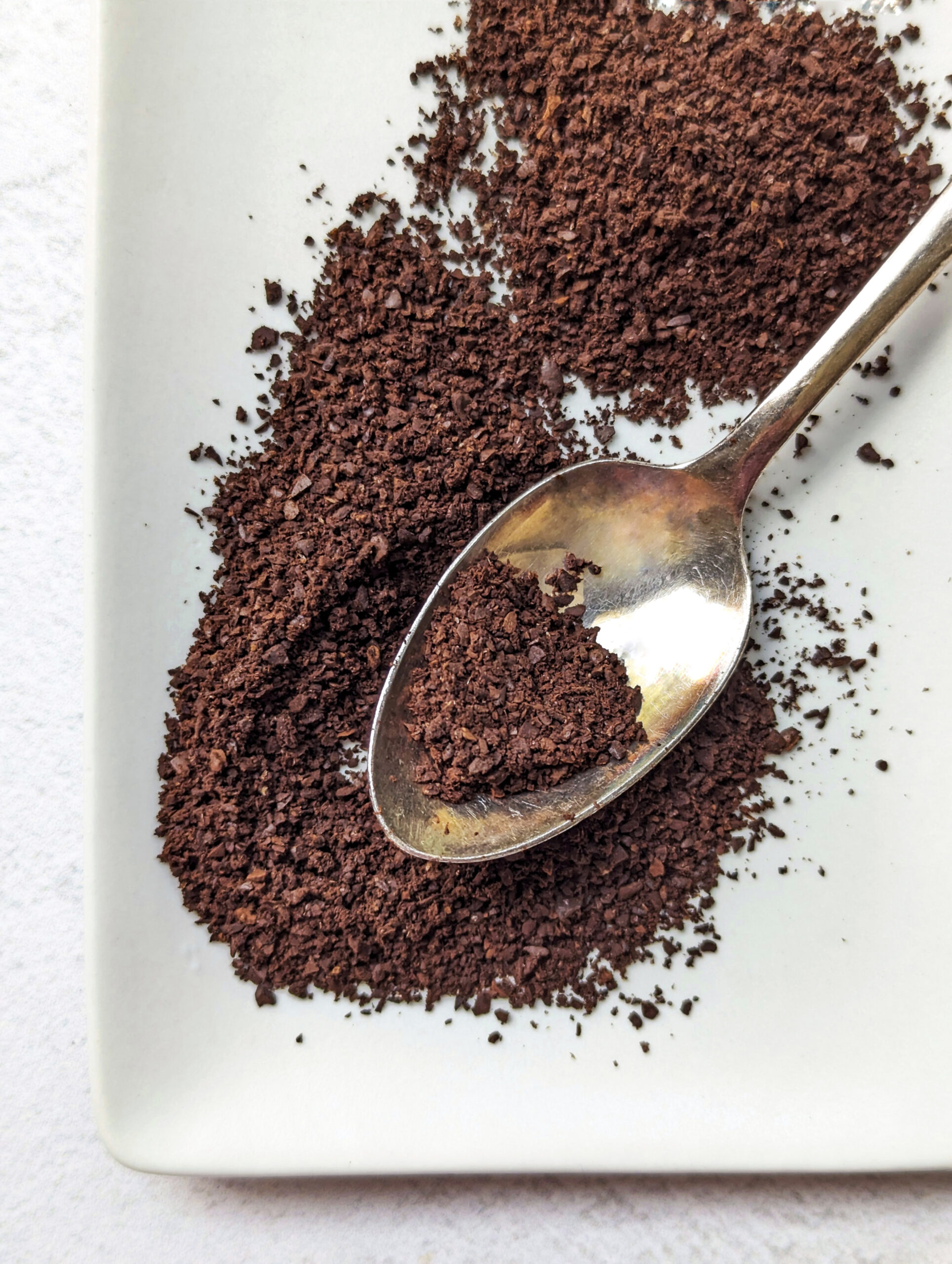 A spoon spreading out coarsely ground coffee beans on a plate.
