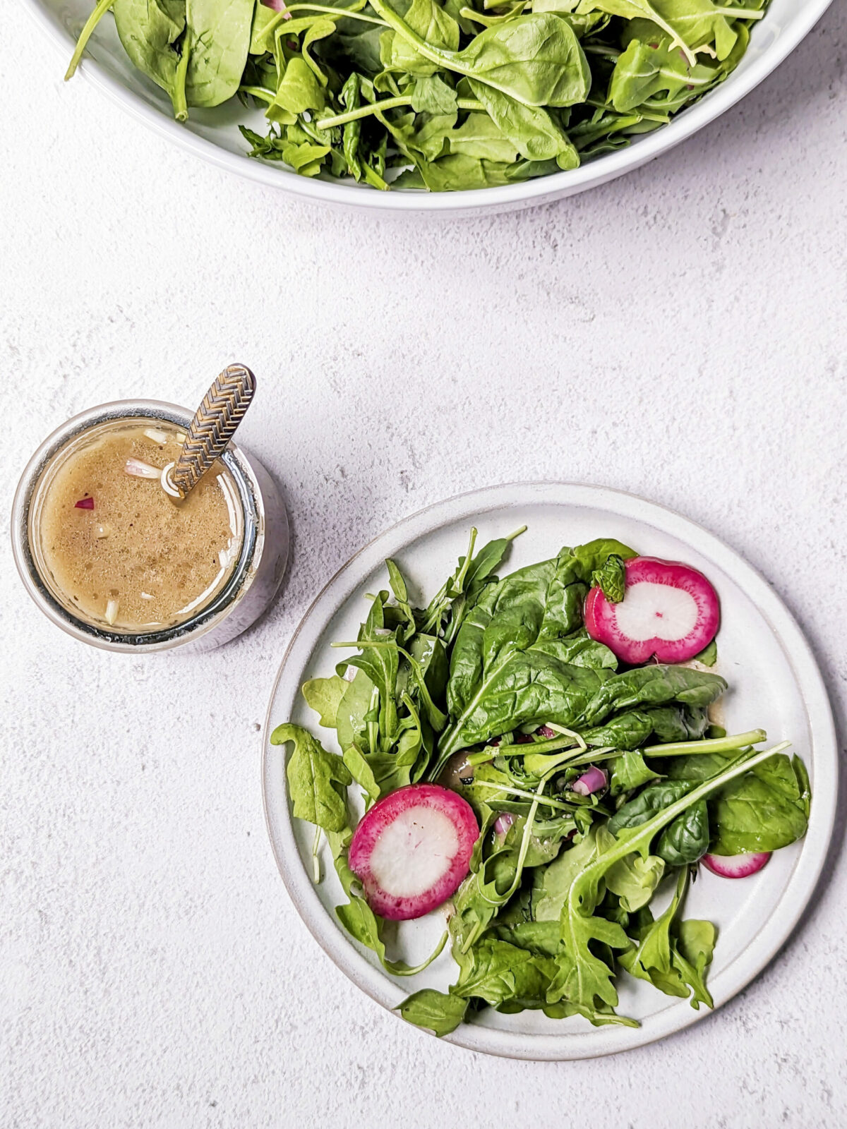 A small plate of Arugula and Spinach Salad with a side of sherry shallot dressing, and a serving bowl of the salad in the background.